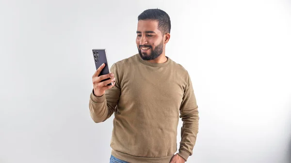 Mutlu genç adam beyaz bir arka planda cep telefonundan içeriğe bakarken gülümsüyor.