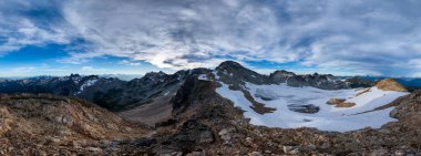 Panoramik manzara kayalık dağları ve parlak gökyüzünün altındaki karlı manzaraları gösteriyor..