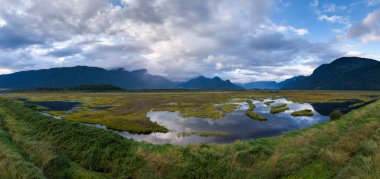 Kanada, BC 'de dramatik bulutlu gökyüzü altında uzak dağları olan güzel bataklıklar..
