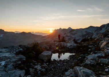 Günbatımında uzak dağları ve yansıtıcı bir havuzu olan kayalık bir tepede yalnız bir yürüyüşçü duruyor. British Columbia, Kanada.