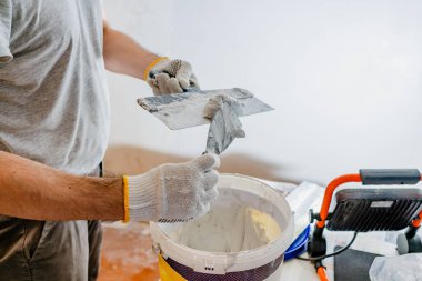 Bir yenileme işçisinin eldivenli elleriyle sıva yapmaya hazırlanırken, daha küçük bir el spatulasından büyük, pürüzsüz bir malayı, daha küçük bir el spatulasıyla malzeme kovasının üzerine yerleştirirken.