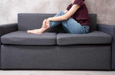 Gri koltukta oturan, mavi kot pantolon ve mor tişörtlü, karın ağrısı ve sindirim sorunları olan bir kadın.