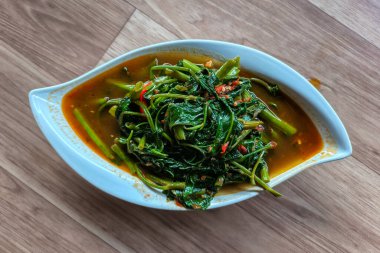 a serving of Kangkung Belacan (stir-fried water spinach with spicy chili shrimp paste) in a white, leaf-shaped bowl on a wooden table.