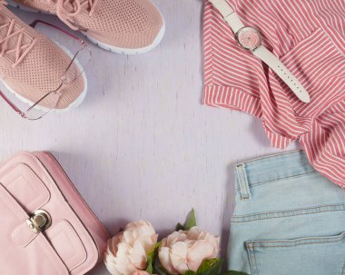 Eşyalar, ahşap arka planda, leylak renginde serilmiş. Küçük pembe el çantası, gömlek, pantolon, gözlük, saat ve açık yaz ayakkabıları. Düzen masanın üzerinde..
