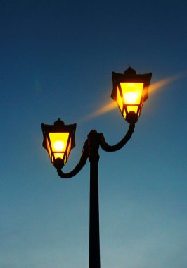 Parkta zarif, klasik bir lamba, akşam ışığında usulca parlıyor. Görüntü, tarihi tasarım ve açık hava aydınlatmasını vurguluyor.