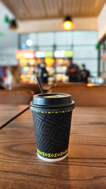 Kahverengi deri sandalyeli ve arka planda insanlar olan bir kafe veya restoranın içi bulanık siyah ve sarı geometrik desenli tek kullanımlık bir kahve fincanı.