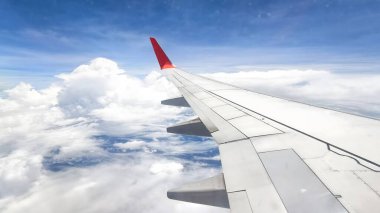 Uçak penceresinden bakıldığında, beyaz bulutlu ve kara bulutlu bir gökyüzüne karşı belirgin kırmızı kanatlı bir uçağın kanadını görüyoruz. peyzaj veya yatay yönelim