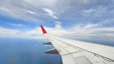 Uçak penceresinden bakıldığında, beyaz bulutlu ve karalı mavi gökyüzüne karşı belirgin kırmızı kanatlı bir uçağın kanadını görüyoruz. peyzaj veya yatay yönelim
