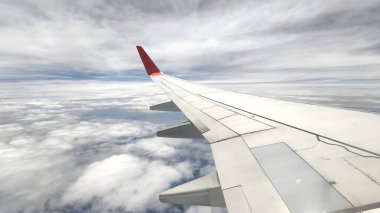 Uçak penceresinden bakıldığında, beyaz bulutlu ve kara bulutlu bir gökyüzüne karşı belirgin kırmızı kanatlı bir uçağın kanadını görüyoruz. peyzaj veya yatay yönelim