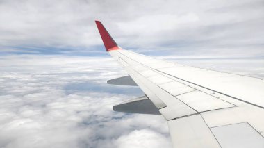 Uçak penceresinden bakıldığında, beyaz bulutlu ve kara bulutlu bir gökyüzüne karşı belirgin kırmızı kanatlı bir uçağın kanadını görüyoruz. peyzaj veya yatay yönelim