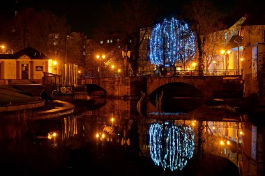 Aydınlatılmış altın çeşme ve arka planında bir katedral kulesi olan karanlık gece. Öndeki altın çağdaş çeşme gecenin karanlığında şehrin ışıklarıyla aydınlanır. Aydınlatılmış tarihi katedralin arka planında bir kule var.. 