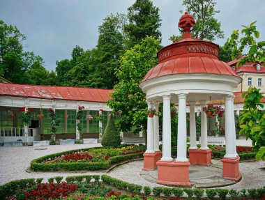 Eski bir kaplıcada çiçekli beyaz bahçe evi. Kırmızı destekli ve çatılı beyaz bahçe evi, çiçekler, yeşil bitkiler ve kaldırımlar etrafta. Arka planda kırmızı çatılı, ağaçlı ve gri gökyüzü olan eski bir spa binası var. Çek Cumhuriyeti