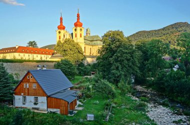 Altında eski bir ev olan güneşle aydınlatılmış tarihi bir kale. İki kuleye sahip sarı şato akşam güneşiyle parlıyor. Kısmen ahşaptan yapılmış eski bir ev, ağaçlar ve nehirle dolu yeşil bir vadide, kalenin altında. Avrupa, Çek Cumhuriyeti