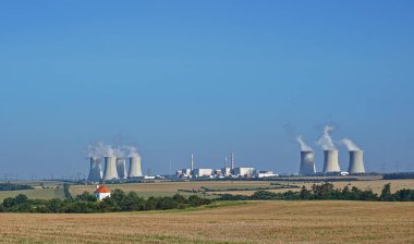 Arka planda nükleer enerji santrali olan kırsal alan. Elektrik santralinde teknik binalar, bacalar ve tüten soğutma kuleleri bulunuyor. Arka planda açık mavi gökyüzü var. Çekçe - Çekçe