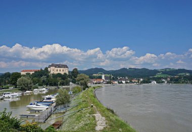 Nehir arasında tersanesi olan körfez. Nehir akışı sağda, küçük tekneler solda körfezde, büyük bir binası olan ağaçlar ve şehir körfezin arkasında, ormanlar, beyaz bulutlar ve mavi gökyüzü arka planda. Tuna Nehri, Avusturya