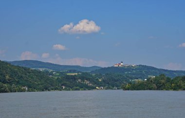 Nehir yüzeyi ön planda, ağaçlı ve tepeli nehir kıyıları arka planda. Kırmızı çatılı beyaz bir kale ve tepenin üstünde iki kule var. Beyaz bulutlu açık mavi gökyüzü tepede, Tuna nehri, Avusturya