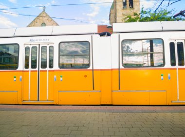 Budapeşte tramvayı yan görüntüsü bir platformda durdu. Görüntü ikonik sarı üniformayı, şehir markasını ve parlak bir gökyüzünün altında bulunan tarihi bir kilisenin kulelerini yakalıyor..