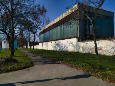 Yeşil cam panelleri ve beyaz bir çevre duvarı olan endüstriyel bir binanın alçak açılı görüntüsü. Kaldırımlar, çıplak ağaçlar ve açık mavi gökyüzünün altındaki derin gölgeler Budapeşte 'nin kentsel havasını yansıtıyor..