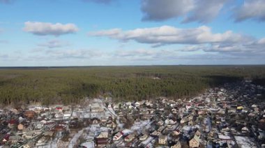 Hava panoraması: İnsansız hava aracı karlı bir yerleşim yerinin üzerinde uçuyor ve bulutlu bir gökyüzünün altında sonsuz bir kozalaklı ormana sorunsuzca yaklaşıyor.. 