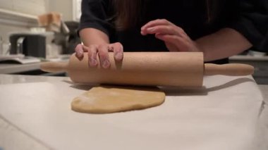Mutfak tezgahında tahta bir oklavayla hamuru yuvarlayan tanınmayan bir kadın. Ev yapımı pastayı sıcak ev ortamında hazırlayan ellerin yakın çekimi.