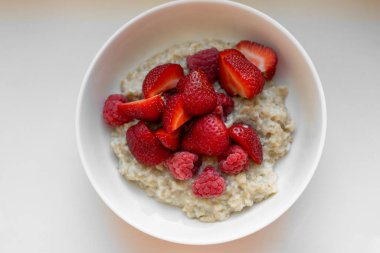 Top-down view of creamy oatmeal served in a white ceramic bowl, topped with halved ripe strawberries and whole raspberries on a white table. The vibrant red berries with the soft beige oats.