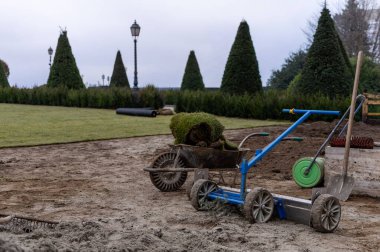 Profesyonel mavi çimenlik döşeme makinesi, yeşil çimenlerin ve çalıların yakınındaki çimlerin üzerinde çim ruloları olan el arabasının yanına yerleştirilmiş çimen halısı için yerleştirilmiş..