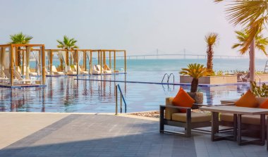 Deniz ve köprü manzaralı tatil köyünde çok güzel lüks bir havuz. Modern tasarım, beyaz şezlong, palmiye ağaçları