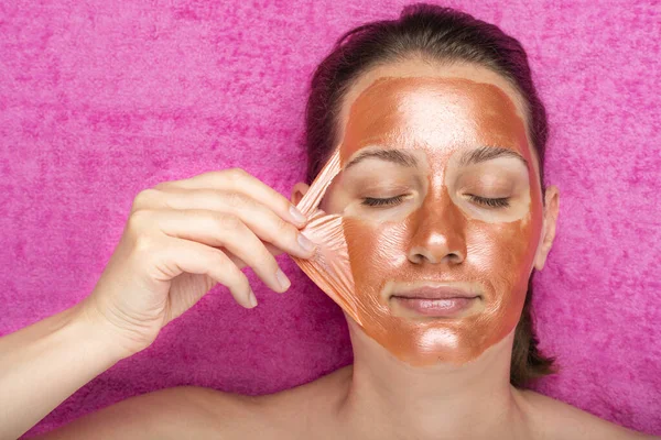 Beautiful girl removes a bronze color cosmetic mask from her face. home facial procedures on a pink background. Facial skin care concept. Copy space