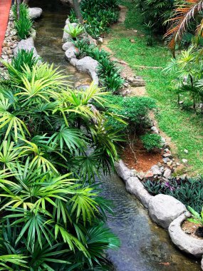 Güneydoğu Asya tarzı bir bahçe, muz yaprakları, palmiyeler ve rattan mobilyalar gibi tropikal bitkilerle doğal elementleri harmanlayarak rahatlatıcı ve sakin bir açık hava ortamı yaratıyor..