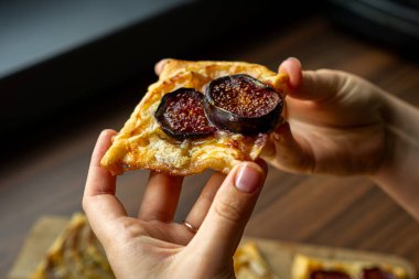 Olgun incir ve erimiş peynirle kaplı bir parça puf böreği tutan bir elin yakın çekimi. Altın kabuk ve zengin meyve dokusu ev yapımı lezzetli bir tatlı atmosferi yaratır. Yemek fotoğrafçılığı, pişirme ve mutfak tasarımı için mükemmel..