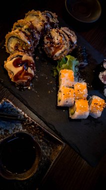 Siyah bir tabakta soslu çok güzel sunulmuş Japon suşi ruloları. Yumuşak ve koyu renkler geleneksel Asya mutfağının zarafetini ve zenginliğini vurgular..