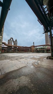 Bulutlu bir gökyüzünün altında paslanmış boruları ve beton yüzeyleri olan eski bir endüstriyel tesisin fotoğrafı. Görüntü simetriyi, çürümeyi ve terk edilmiş bir bitkinin atmosferini yakalar. Endüstri, mimari ve kentsel keşifler hakkında konular için ideal.