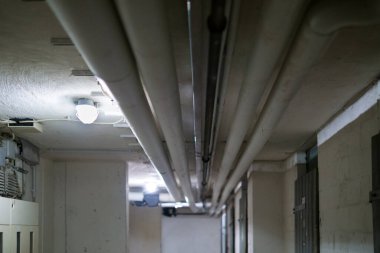Lambalarla aydınlatılmış bodrum tavanı boyunca uzanan boruların ve kanalların görüntüsü. Görüntü endüstriyel altyapıyı ve mühendislik, bakım ve yapı sistemlerini temsil eden bir servis koridorunun karmaşık teknik düzenini vurguluyor.