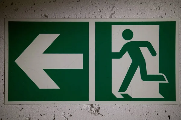 Sol tarafı ok olan bir duvarda yeşil bir acil çıkış tabelasının yakın çekimi. Görüntü, kamusal ve endüstriyel binalarda kaçış yollarını belirlemek için kullanılan güvenlik, tahliye ve rehberliği temsil ediyor..