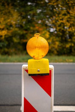 Kırmızı ve beyaz bir trafik barikatına asılmış parlak sarı uyarı ışığı bir yol inşaatı alanında duruyor. Görüntü, altyapı, ulaştırma ve kamu güvenliği temalarının resmedilmesi için uygun güvenlik, ihtiyat ve kentsel bakım sunmaktadır.