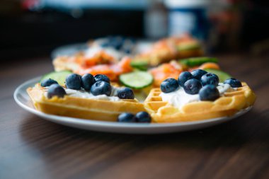 Makro yakın plan çekimde üzerinde krema ve taze yaban mersini olan altın waffle 'lar gösteriliyor. Detaylı görünüm, gevrek waffle dokusu ile yumuşak meyve arasındaki zıtlığı vurgular. Yemek tatlı, canlı ve görsel olarak zengin görünüyor..