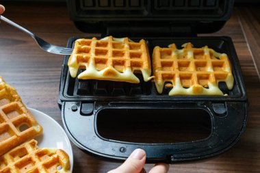 Bir çatal taze pişmiş waffle 'ı kaldırır ve altın kabuğunu ve yumuşak dokusunu ortaya çıkarır. Görüntü sıcak bir ev yapımı yemek anı gösteriyor. Izgara deseni imzalı waffle şeklini vurgular.