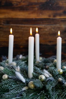 Dört uzun beyaz mum, altın süslemelerle süslenmiş donmuş yeşil bir çelengin üzerinde parlak bir şekilde yanar. Sıcak alevler, sade ahşap arka planla çok güzel tezat oluşturuyor. Sıcak ve şenlikli bir tatil ortamı yaratıyor..