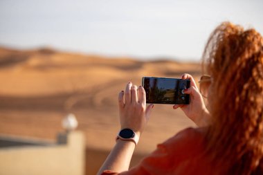 Kızıl saçlı bir kadın, Rub al Khali çölündeki dalgalı kum tepecikleri manzaralı çitlerle çevrili bir bahçenin fotoğrafını çekiyor. Yüksek kalite fotoğraf