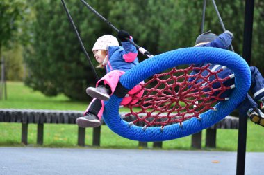 Çocuklar sonbahar parkında salıncakta sallanıyor. Su geçirmez takım elbiseli neşeli kardeşler dışarıda oyun saatlerinde gülüşür ve hareket ederler. Erkek ve kız kardeş oyun alanında birlikte eğleniyorlar..