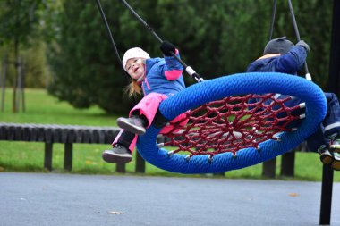 Çocuklar sonbahar parkında salıncakta sallanıyor. Su geçirmez takım elbiseli neşeli kardeşler dışarıda oyun saatlerinde gülüşür ve hareket ederler. Erkek ve kız kardeş oyun alanında birlikte eğleniyorlar..