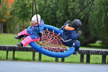 Çocuklar sonbahar parkında salıncakta sallanıyor. Su geçirmez takım elbiseli neşeli kardeşler dışarıda oyun saatlerinde gülüşür ve hareket ederler. Erkek ve kız kardeş oyun alanında birlikte eğleniyorlar..