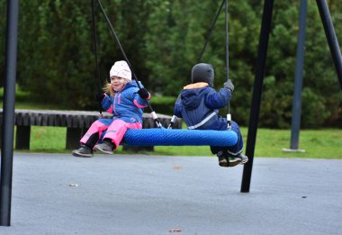 Çocuklar sonbahar parkında salıncakta sallanıyor. Su geçirmez takım elbiseli neşeli kardeşler dışarıda oyun saatlerinde gülüşür ve hareket ederler. Erkek ve kız kardeş oyun alanında birlikte eğleniyorlar..