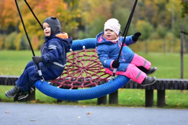 Sonbahar parkında yuvarlak bir salıncakta sallanan iki çocuk. Renkli su geçirmez tulumlar içinde mutlu erkek ve kız kardeşler serin bir sonbahar gününde dışarıda oyun oynamanın keyfini çıkarıyorlar. Kardeşler birlikte eğleniyor.