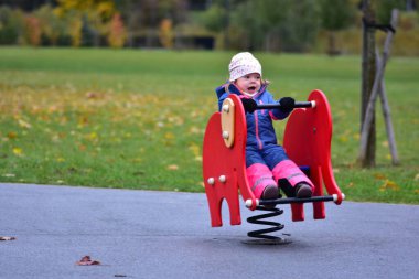 Gülen küçük kız yeşil bir parkta bahar oyuncağına biniyor. Parlak yağmurluk giymiş heyecanlı çocuk, sonbahar yaprakları ve serin havayla çevrili açık havada eğleniyor..