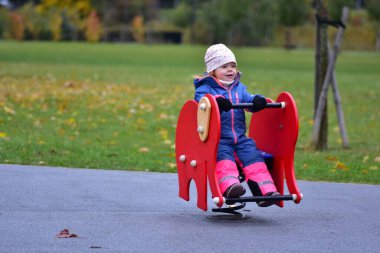 Gülen küçük kız yeşil bir parkta bahar oyuncağına biniyor. Parlak yağmurluk giymiş heyecanlı çocuk, sonbahar yaprakları ve serin havayla çevrili açık havada eğleniyor..
