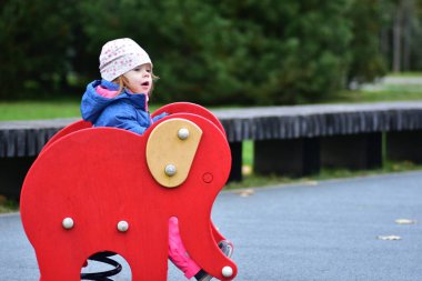 Küçük kız kırmızı bir fil bahar oyuncağının üzerinde oturuyor. Sıcak ceketli ve şapkalı şirin çocuk serin bir sonbahar sabahında oyun oynamaktan hoşlanıyor..