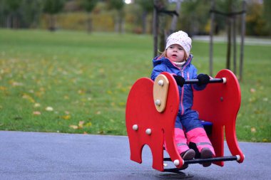 Gülen küçük kız yeşil bir parkta bahar oyuncağına biniyor. Parlak yağmurluk giymiş heyecanlı çocuk, sonbahar yaprakları ve serin havayla çevrili açık havada eğleniyor..