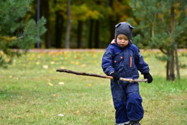 Sıcak mavi tulumlu gülen çocuk, sonbahar parkında dışarıda oynarken elinde sopa tutuyor. Mutlu çocuk ağaçlar ve düşen yapraklar arasında doğayı keşfetmekten hoşlanıyor..