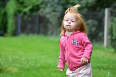 Pembe kapüşonlu, neşeli bir ifadeyle yeşil çimlerin üzerinde koşan, açık havada eğlenen ve açık havada bahçede temiz hava alan neşeli kızıl saçlı bir çocuk..